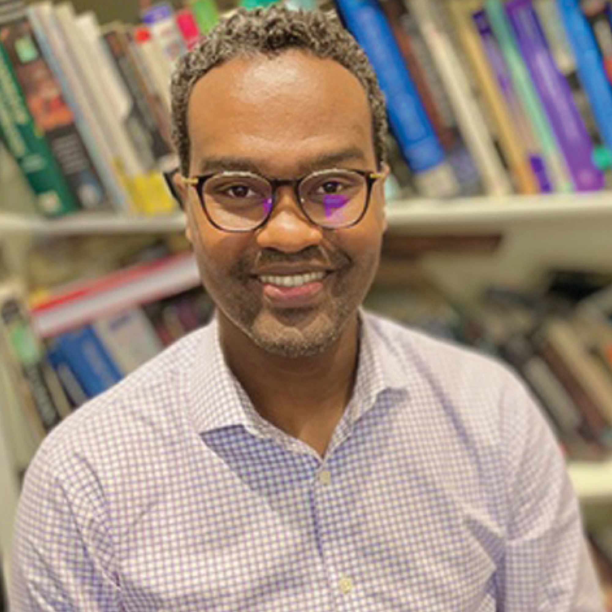 A brown-skinned man with short hair and a light beard in glasses and a button up shirt smiles for a portrait in front of a bookshelf