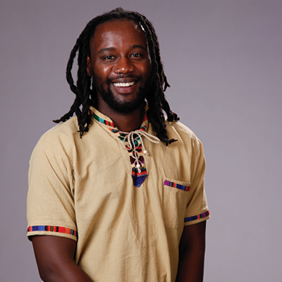 A black man with long hair in a shirt with colorful trim smiles for a portrait in front of a gray background