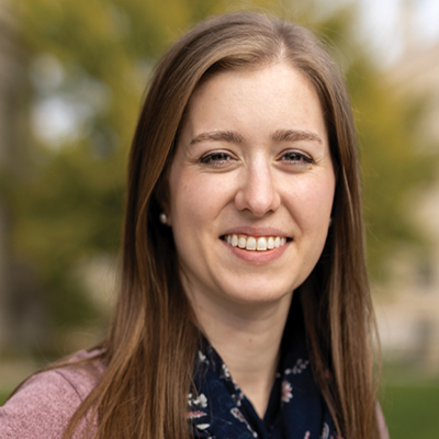 A white woman with straight brown hair in a black scarf and purple shirt smiles for a portrait in front of blurred trees