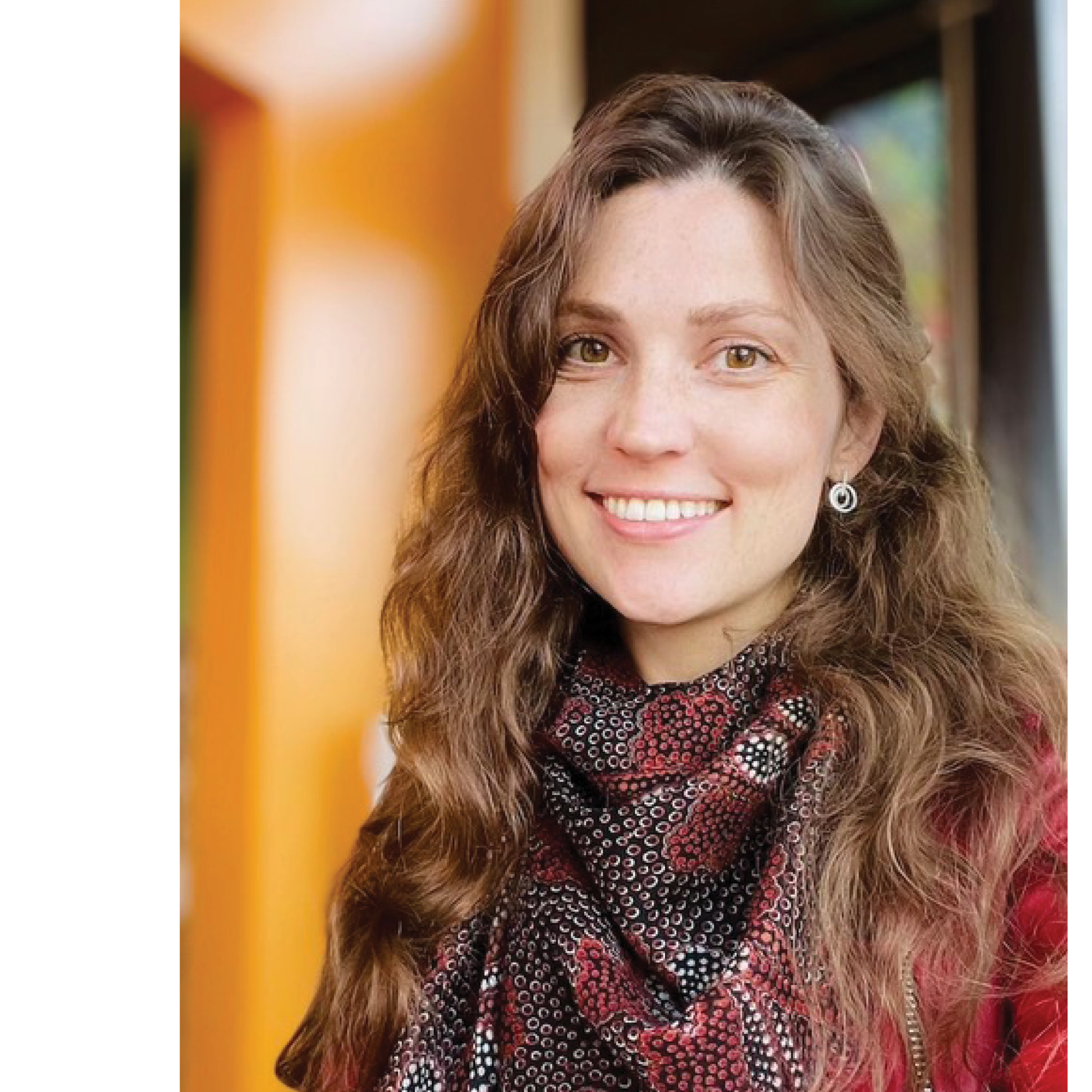 A woman with wavy brown hair in a scarf smiles for a portrait in a wooden room