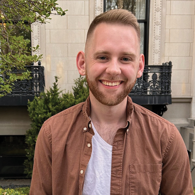 A white man with short hair and a short beard in a brown shirt smiles for a portrait outside of a stone house