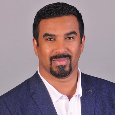 A middle-eastern man with dark hair and beard in a blue suit smiles for a portrait in front of a grey background